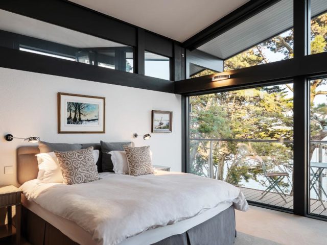 Huf Haus ART Sonder - Doppelbett mit Tagesdecke und Kissen vor Panoramafenstern mit Blick aufs Meer