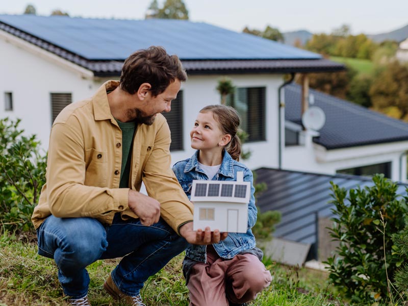 Kleines Mädchen und ihr Vater halten ein Paiermodell eines Hauses mit Solarpaneelen auf dem Dach.