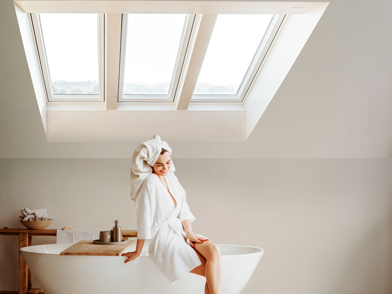 Eine Frau sitzt im Bademantel und mit Handtuchturban auf dem Rand einer freistehenden Badewanne unter drei Dachflächenfenstern