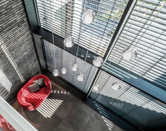 Wohnbereich mit darüberliegendem Luftraum indem ein kleines rotes Sofa steht und außenliegenden Jalousien als Schattenspender