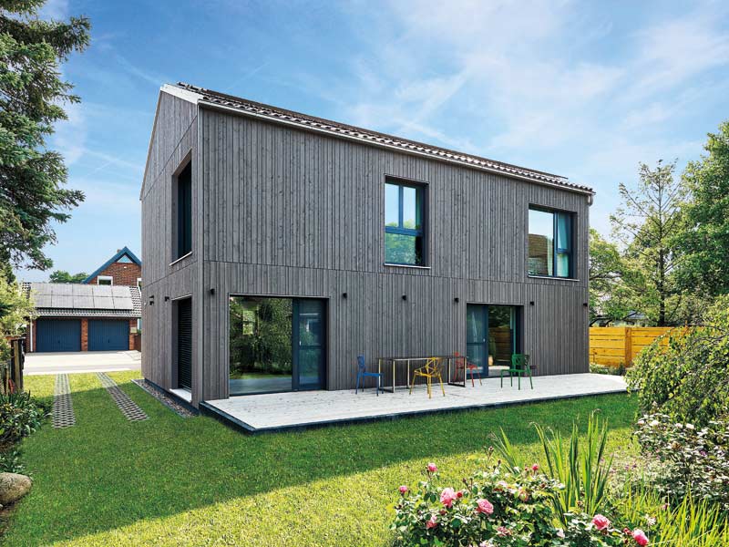 my smart green home individuell von Baufritz Aussenansicht zum Garten mit heller Holzterrasse, die sich abhebt von der dunklen Holzfassade