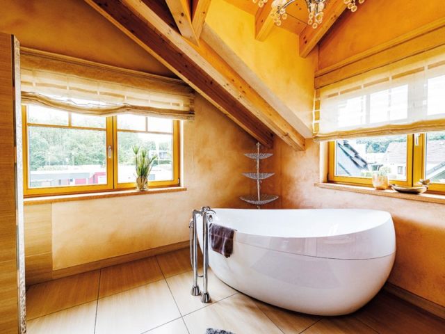 Elegante weiße freistehende Badewanne in einem Holzhaus zwischen zwei Fenstern und mit freistehender Armatur davor.