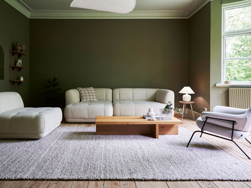 Wohnbereich in einem Altbau mit grünen Wänden, hellem Sofa, Sessel und Stuhl mit kleinem Holztisch auf einem grauen Teppich.