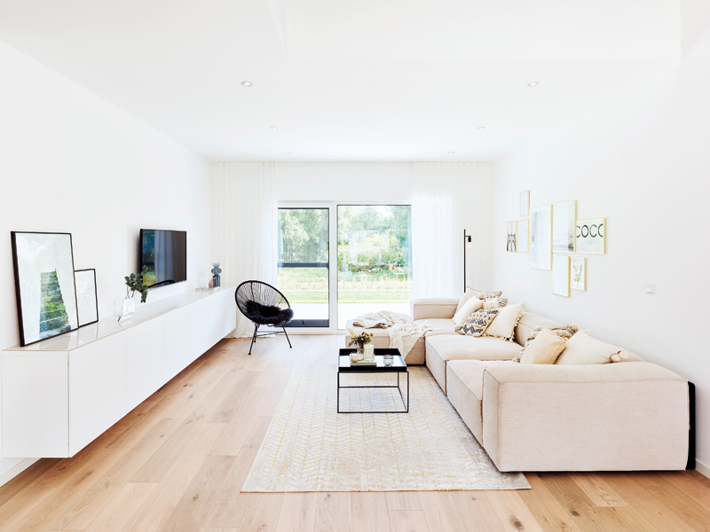 Fertighaus Weiss Blick vom Essplatz in den Wohnbereich, an rechter Wand langes weißes Ecksofa, davor flaches Tischchen, gegenüber schmales weißes Sideboard, zum Sofa passender heller Teppich auf den Holzdielen, schwarzer Korbsessel vor bodentiefer Glastür