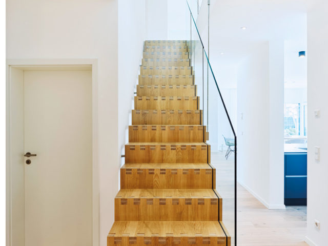 Haus Rohr von Fertighaus Weiss; gerade massive Holztreppe mit Glasgeländer im hellen Flur, rechts Blick in die Küche, links Zimmertür zum Arbeitszimmer