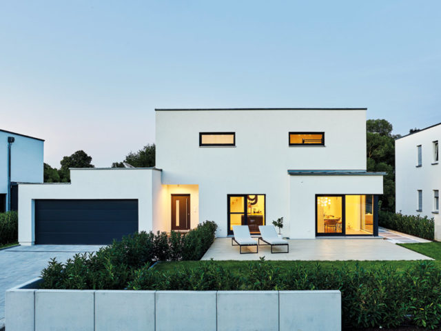 Der Hauseingang dieser weißen Baushausvilla ist als Gebäuderücksprung gestaltet. Diese architektonische Entscheidung akzentuiert die Fassade und bietet wirkungsvollen Wetterschutz; links im Bild große Garage, am Fuß des Bildes Hecke teilweise von Betonzaunteilen verdeckt vor kleiner Rasengläche