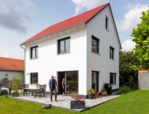 Haus Space 4 XL der Deutschen Hausmanufaktur, Außenansicht zweigeschossiges und weiß verputztes Satteldachhaus; Rückfront zum Garten hin mit direktem Zugang, auf möblierter Terrasse Baupaar mit Hund