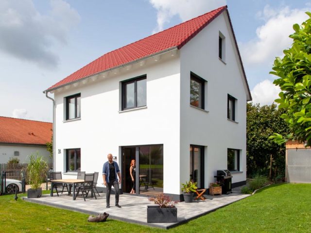 Haus Space 4 XL der Deutschen Hausmanufaktur, Außenansicht zweigeschossiges und weiß verputztes Satteldachhaus; Rückfront zum Garten hin mit direktem Zugang, auf möblierter Terrasse Baupaar mit Hund