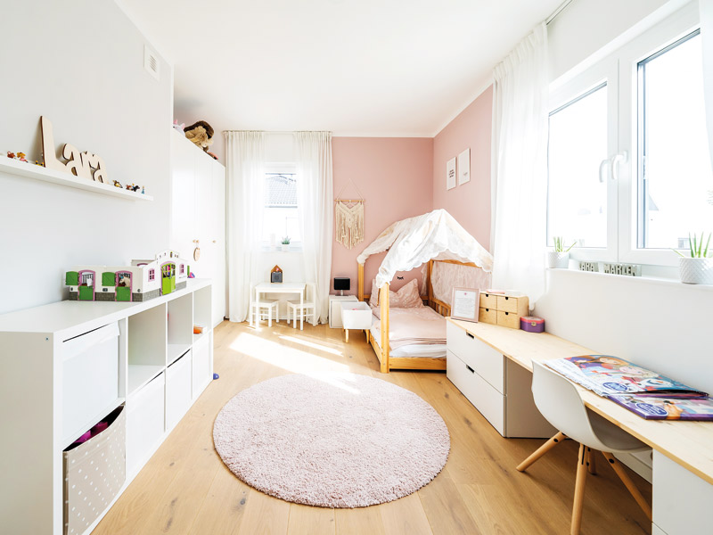 Modernes Kubushaus von Streif Haus, Kinderzimmer, links im Bild weißes teilweise offenes Regal, an der Rückwand Kindertisch mit Hockern und Himmelbett, auf der rechten Seite unter dem Fenster lange Schreibplatte mit Stuhl, als Seitenwände dienen breite Regale mit Schubkästen, heller Dielenboden mit flauschigem runden Teppich