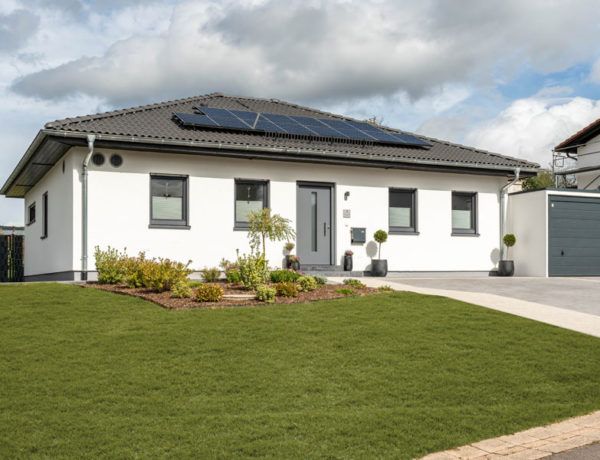 Bungalow mit Doppelgarage von Streif Haus, Vorderansicht mit grauer Haustür, vier kleineren Fenstern und dem anthrazitfarbenen Dach mit PV-Anlage. Rechts geht es zu den Garagen. Im Vordergrund Wiese, links neben der Haustür ein kleiner Vorgarten