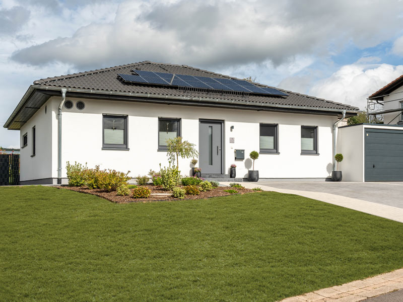 Bungalow mit Doppelgarage von Streif Haus, Vorderansicht mit grauer Haustür, vier kleineren Fenstern und dem anthrazitfarbenen Dach mit PV-Anlage. Rechts geht es zu den Garagen. Im Vordergrund Wiese, links neben der Haustür ein kleiner Vorgarten