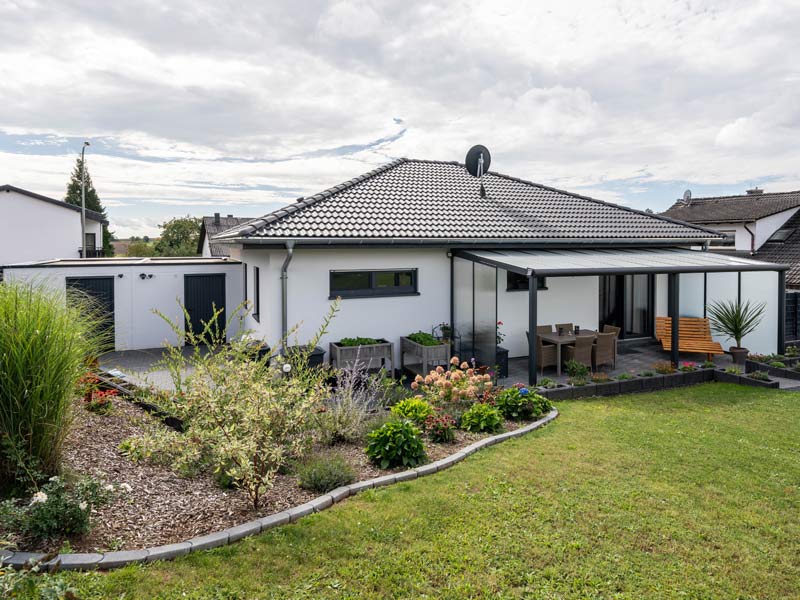 Bungalow mit Doppelgarage von Streif Haus, Rückansicht mit überdachten möblierter Terrasse und Rückansicht der Garage, direkter Übergang zum Garten mit Wiese und Beet mit Büschen und Sträuchern