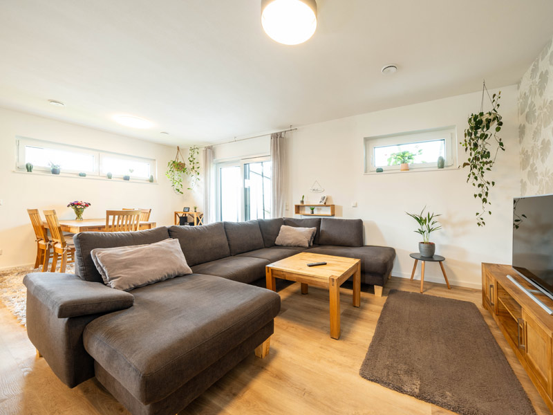 Bungalow mit Doppelgarage von Streif Haus, Blick ins Wohnzimmer mit hellen Holzdielen, im Vordergrund dunkrlbraune Sitzlandschaft mit flachem Tisch und Sideboard mit TV-Gerät, im Hntergrund Esstisch aus Holz und Holzstühle, schmale Fensterbänder an zwei Wänden und eine bodentiefe Fenstertür sorgen für Licht