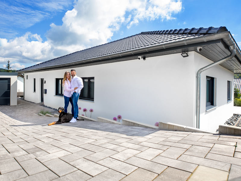 Bungalow Vintra von Fingerhut Haus, Vorderansicht des weiß verputzten Bungalows mit anthrazitfarbenem Wanlmdach mit Eingangstür und zwei kleineren Fenstern, davor das Baupaar mit Hund, Einfahrt mit großen hellen Platten versehen, links angeschnitten die ebenfalls weiß verputzte Garage