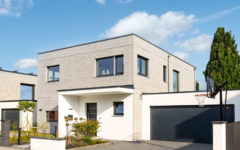 Entwurf Kirschner von SchwörerHaus Straßenansicht, beige geklinkerter zweistöckiger Bauauswürfell mit angeschlossener Garage, überdachter Hauseingang und Garage weiß verputzt, kleriner begrünter Vorgarten, gepflasterter Garagenvorplatz, Baum und Basketballkorb, Übergang zur Straße ohne Zaun