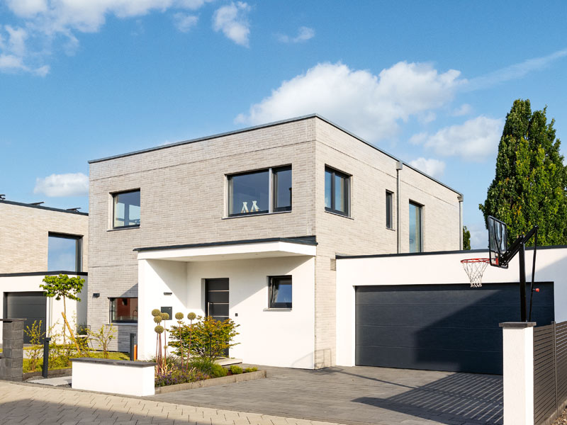 Entwurf Kirschner von Schwörerhaus Straßenansicht, beige geklinkerter zweistöckiger Bauauswürfell mit angeschlossener Garage, überdachter Hauseingang und Garage weiß verputzt, kleriner begrünter Vorgarten, gepflasterter Garagenvorplatz, Baum und Basketballkorb, Übergang zur Straße ohne Zaun