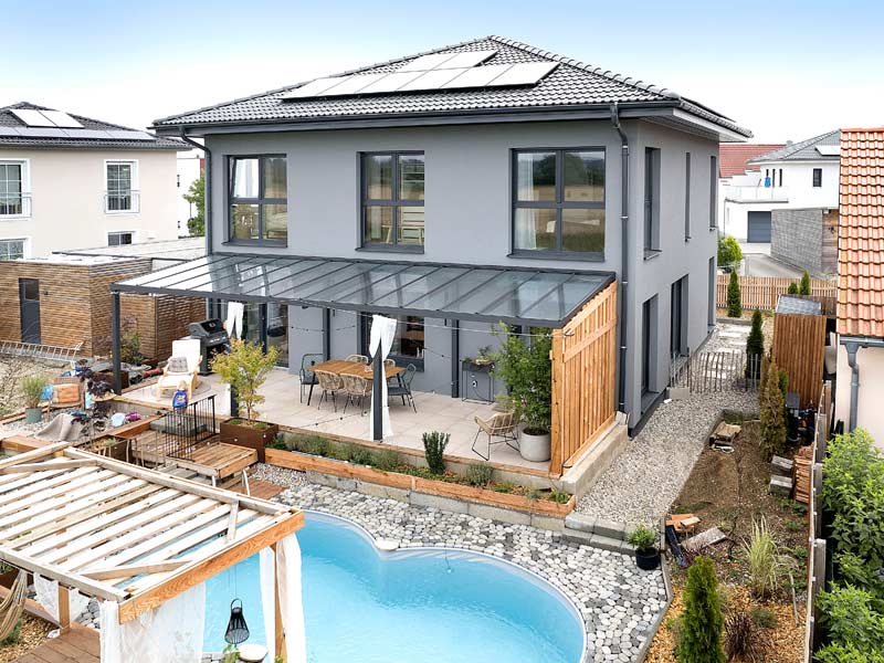 Moderne zweigeschossige Stadtvilla mit grauer Putzfassade, großen Fenstern, Satteldach mit Photovoltaikanlage, großer mit Glas überdachter Terrasse und Pool im Garten.