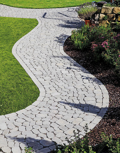 Gepflasterter Gartenweg mit geschwungenem Verlauf, umgeben von Rasen, Blumenbeeten und einer Steinmauer im Hintergrund.