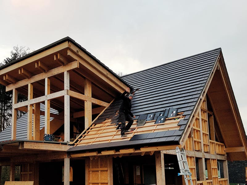 Auf dem Dach eines neu errichteten Hauses steht ein Dachdecker und verlegt Solarziegel für die Produktion von Solarstrom.