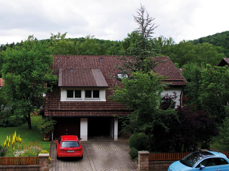 Älteres Einfamilienhaus umgeben von viel Baumbestand im Garten und zwei Fahrzeugen davor stehend.