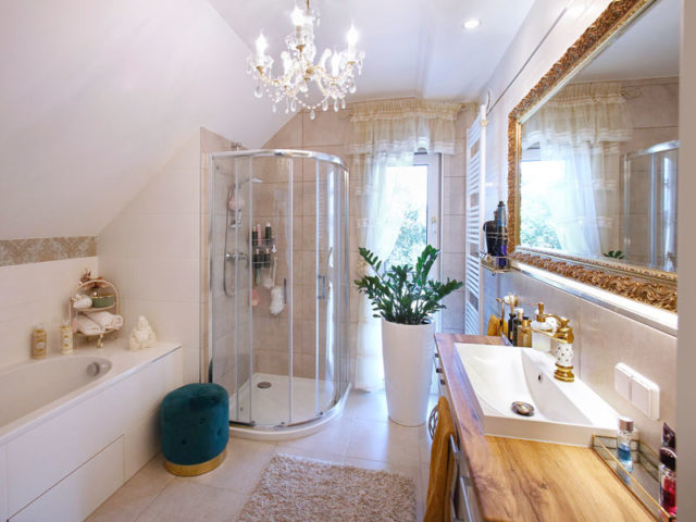 Badezimmer mit eingefliester Badewanne, Eckdusche, großem Spiegel mit goldener Umrandung, Waschtisch aus Holz, Kronleuchter und Pflanze in einem weißen Topf.