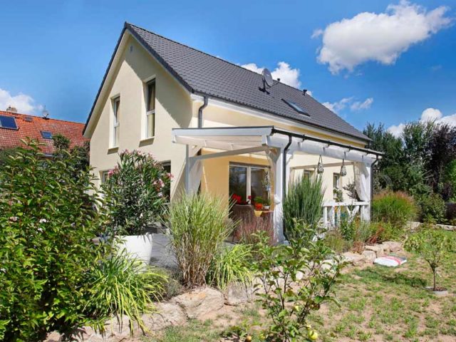 Danwood Family 133 V1: Haus mit gelber Fassade, Satteldach, überdachter Terrasse und gepflegtem Garten mit Büschen und Pflanzen.