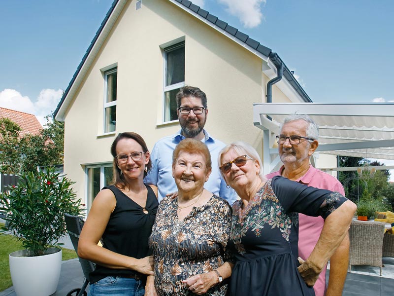 Drei Generationen einer Familie stehen vor einem gelben Satteldachhaus und lächeln zufrieden.