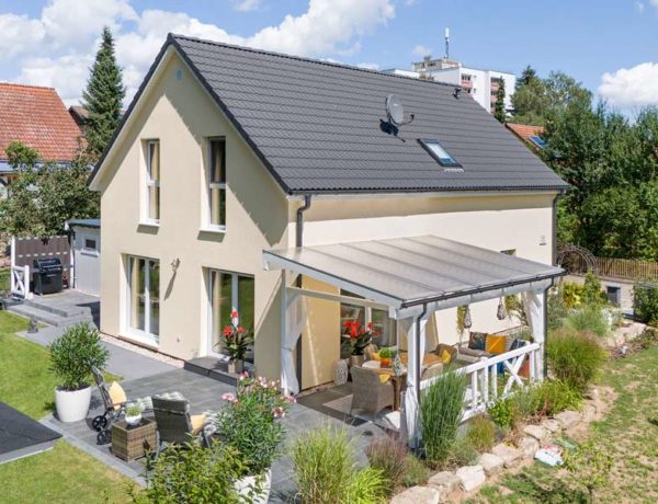 Danwood Family V133 V1: modernes Satteldachhaus mit gelber Putzfassade und weißen Fenstern und großer teilweise überdachter Terrasse mit Sitzmöglichkeiten