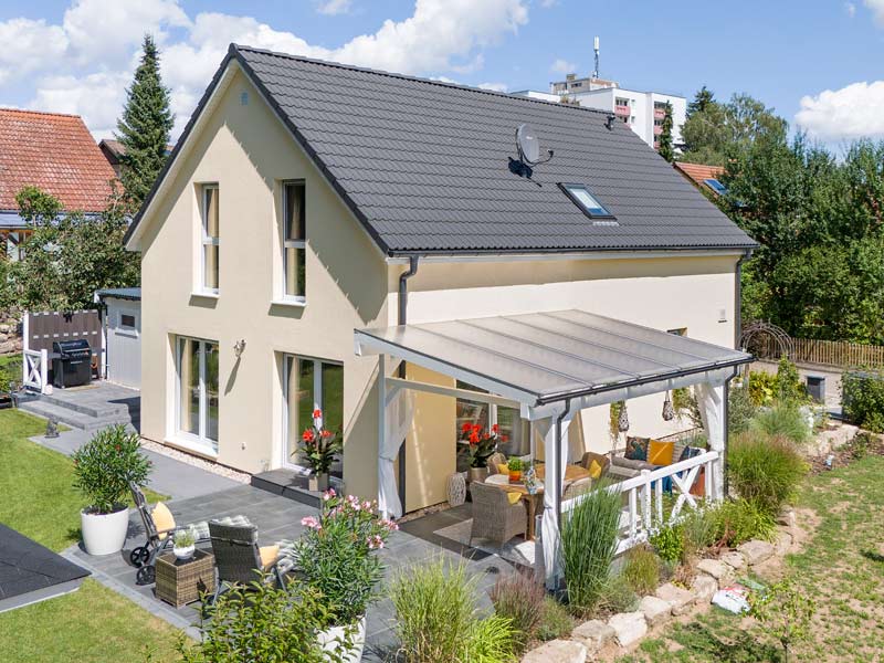 Danwood Family V133 V1: modernes Satteldachhaus mit gelber Putzfassade und weißen Fenstern und großer teilweise überdachter Terrasse mit Sitzmöglichkeiten